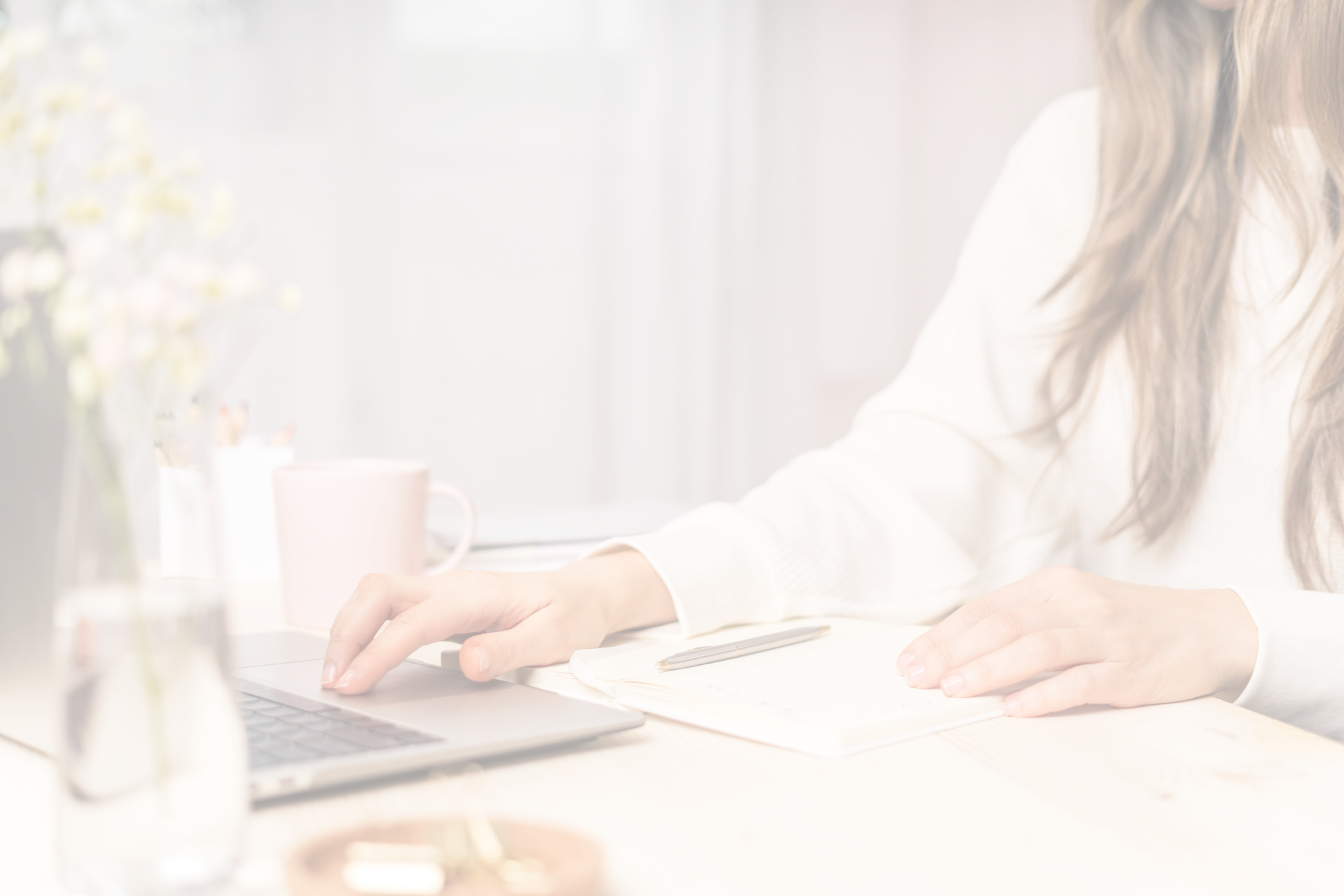 A woman with light brown hair and a white shirt working on a laptop computer. A coffee cup, flowers, and a notebook sit next to her laptop.